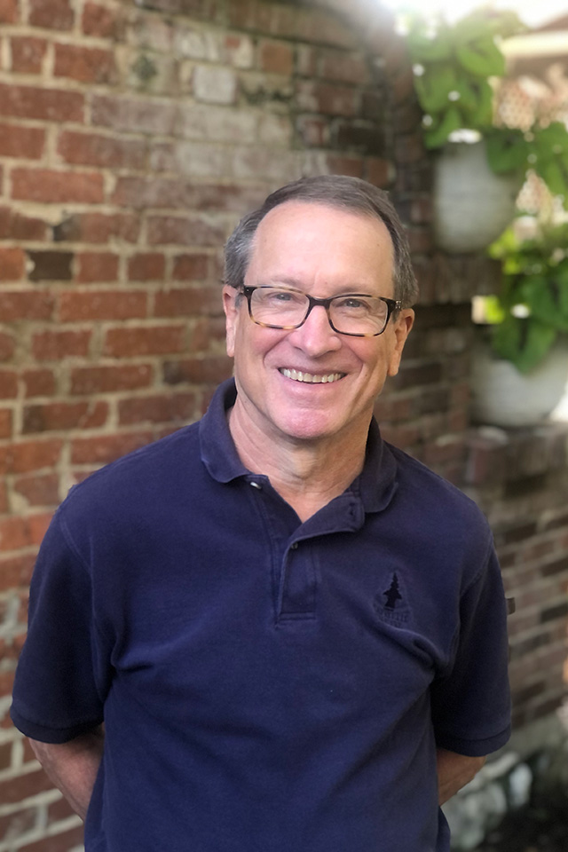 A man poses for a photo with a brick wall and plants behind him.