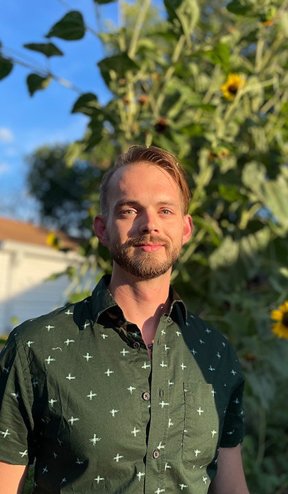 Geoffrey Brewer stands outdoors in front of a tree, wearing a green shirt.