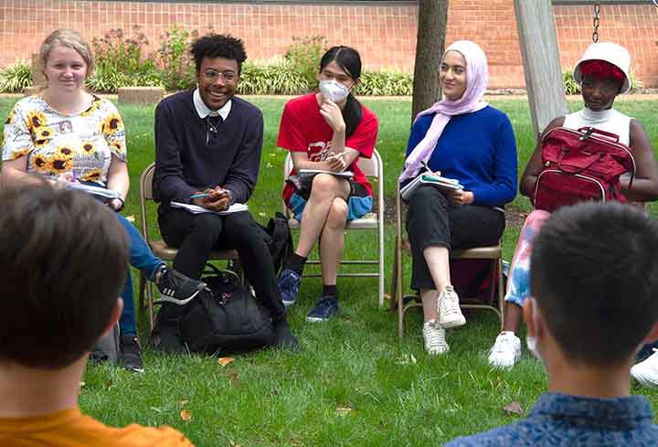 A group of students sit on folding chairs set up in a circle on a lawn for discussion