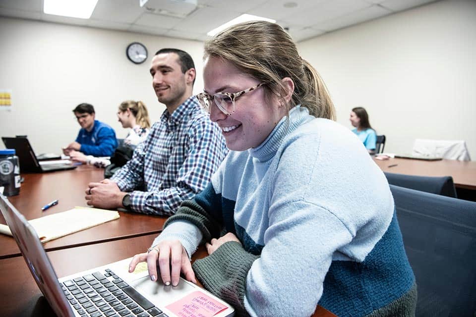 A student wearing glasses and a sweater works on her laptop in a graduate education course