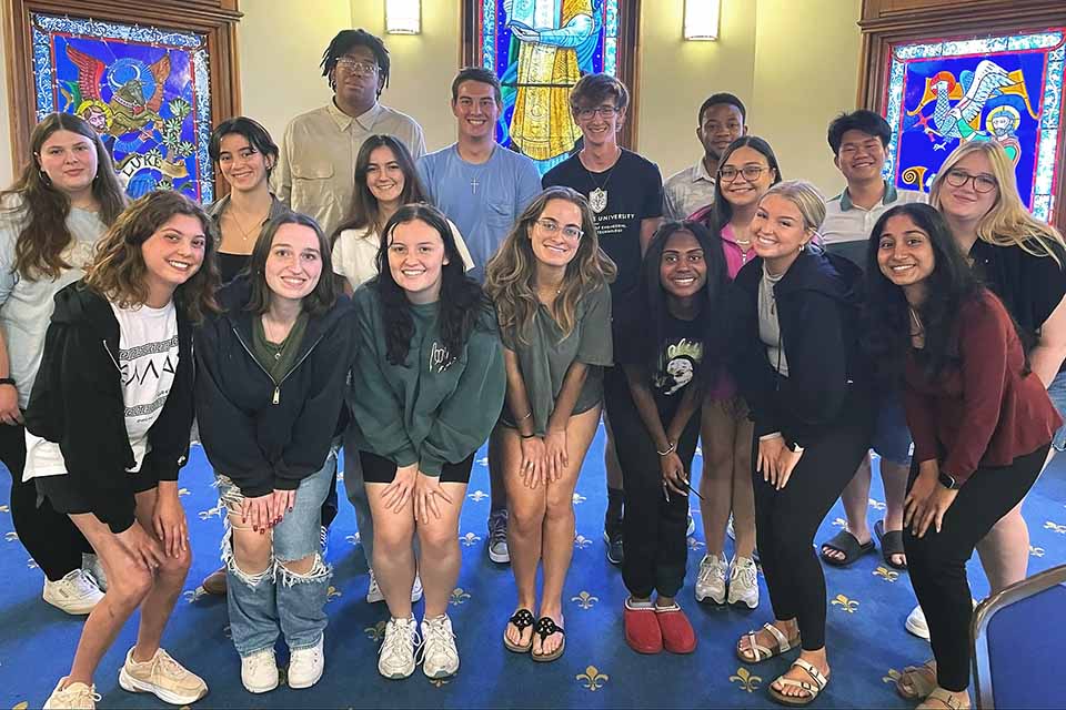 A group of 17 students pose for a photo together in a room with stained glass windows behind them.