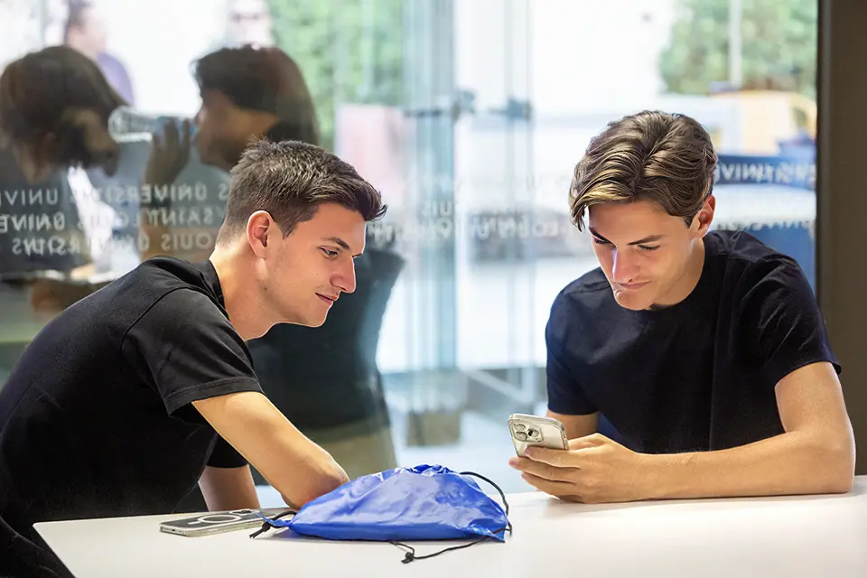 Two students converse while sitting at a table in an area on campus. One student looks at his phone.