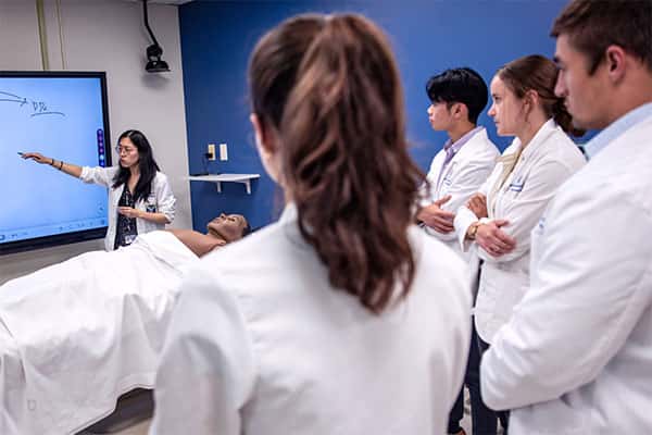 An instructor presents to a class of students in white coats in the Clinical Skills Lab.