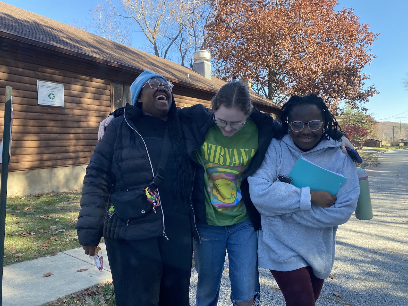 Three Micah students laughing as they walk outdoors with a blue sky and a log building in the background