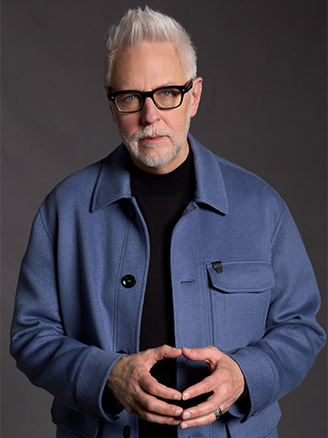 James Gunn, with short, spiked white hair and glasses, wearing a blue jacket over a black shirt, poses thoughtfully with his hands loosely clasped against a dark gray background. James Gunn, with short, spiked white hair and glasses, wearing a blue jacket over a black shirt, poses thoughtfully with his hands loosely clasped against a dark gray background.