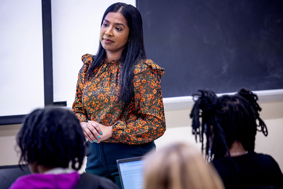 Professor Popy Begum, Ph.D., wearing a floral blouse, leads a classroom discussion. Students, some with laptops, focus on her.