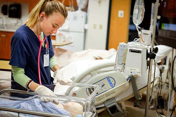 A female student, dressed in scrubs, uses a stethoscope to examine an infant manikin lying in a bassinet. Another hospital bed can be seen in the background.