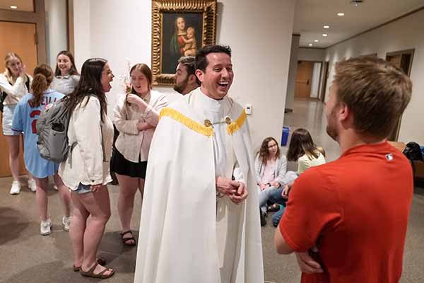 A man wearing white vestments speaks to a student. Other students can be seen chatting in the background.