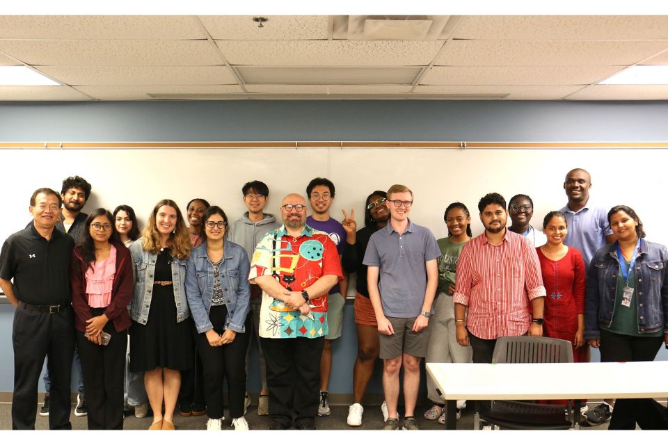 A group of approximately 20 students, faculty and staff interested in maternal child health stand, posing in front of a white board in a classroom. 