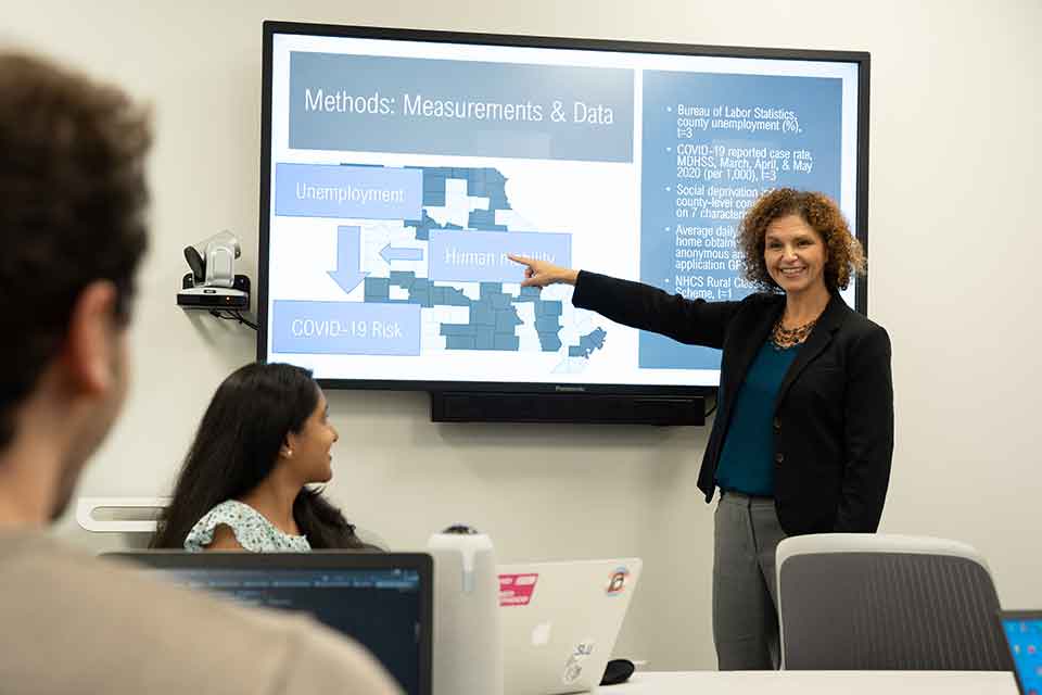A woman points to a TV screen that is displaying data from a recent study. Two students look on. 