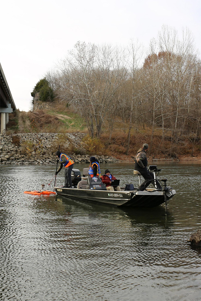 Four people are on a boat in a river, working with various pieces of equipment. Behind them, a shoreline with rocks and trees without leaves are visible.